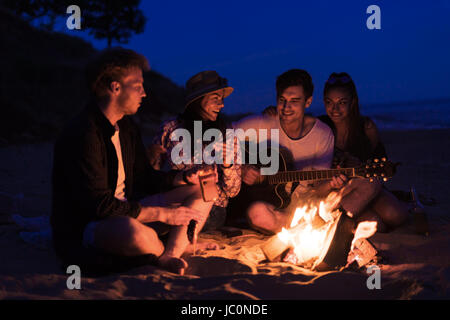 friends sitting on the beach. man is playing guitar. Stock Photo