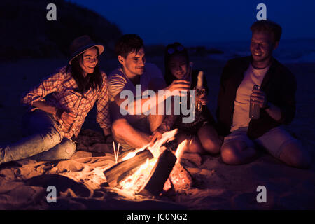 friends sitting on the beach clink glasses near bonfire Stock Photo