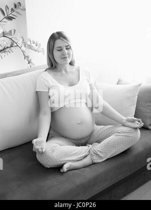 Black and white photo of young pregnant woman exercising on sofa at living room Stock Photo