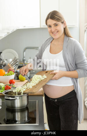 Cute smiling pregnant woman putting vegetables in soup Stock Photo