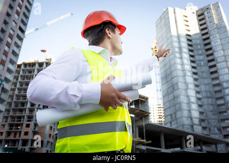 Closeup portrait of foreman with blueprints pointing hand at high building Stock Photo