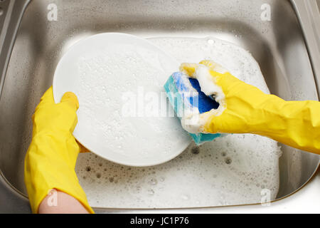 Close up of female hands in yellow protective rubber gloves washing white plate with blue cleaning sponge against kitchen sink full of foam Stock Photo