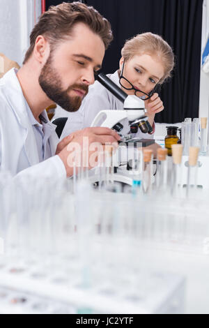 Little girl student with eyeglasses looking at teacher working with microscope in lab Stock Photo