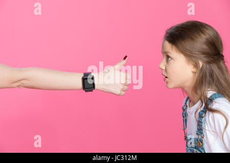 Side view of shocked little girl looking at woman with smartwatch showing thumb up Stock Photo