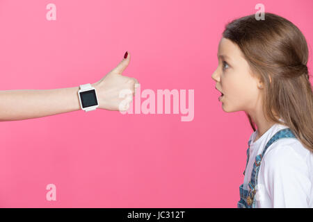 Side view of shocked little girl looking at woman with smartwatch showing thumb up Stock Photo