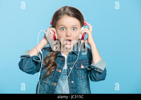 Shocked little girl wearing headphones holding smartphone and looking at camera Stock Photo