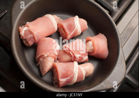 Cooking pigs in blankets, or rashers of bacon wrapped around beef and pork sausages, with a batch of uncooked piggies in a cast iron casserole dish waiting to be cooked in the oven or on a hot griddle Stock Photo