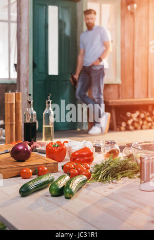 Close-up view of wooden table with meat and vegetables for bbq and man with beer on porch Stock Photo