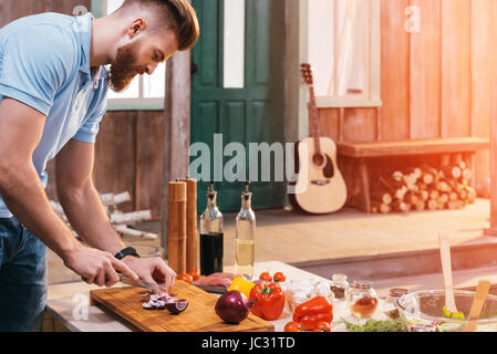 Side view of bearded young man cutting onion for barbecue Stock Photo