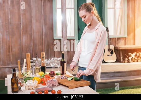Smiling young woman cutting cucumber at table with meat and vegetables outdoors Stock Photo