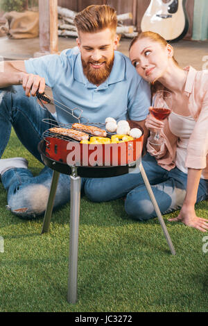 man and woman roasting meat and vegetables on barbecue grill, woman holding wineglass Stock Photo