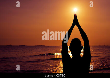 the silhouette of a young caucasian man practicing yoga in front of the sea in backlight Stock Photo