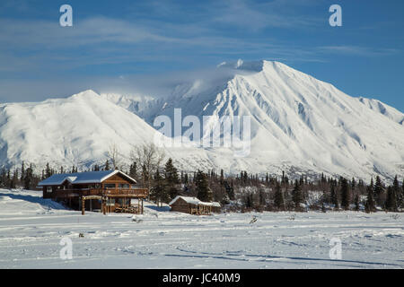 Log cabins in Alaska sit at the base of a frozen lake with snowy mountains in the background. Stock Photo