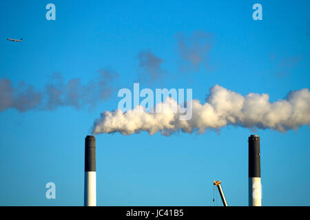 A jet flies over a factory with smokestacks puffing smoke. Amager Bakke waste-to-power incinerator plant aims to become the worldâs first zero-carbon city by 2025. There are also plans to make an artificial ski slopw and outdoor climbing route off the top and up the face of the power plant. The power plant burns garbage but Danish residents are so adept at recycling that garbage often needs to be imported from nearby countries. Stock Photo