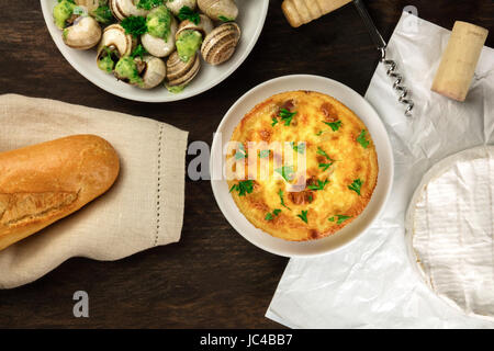 An overhead photo of an assortment of typical French foods on a dark rustic background texture with a place for text. Baguette, snails in green sauce, Stock Photo