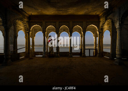 A indian woman is standing on a illuminated balcony with beautifully carved pillars at night, overlooking the holy river Ganges Stock Photo