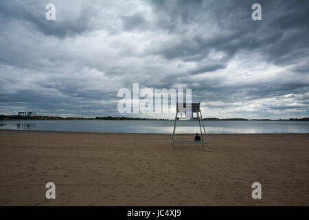 One person sits huddled near the lifeguard stand on the beach near Castle Island, South Boston, MA. Causeway surrounding Pleasure Bay is visible. Stock Photo