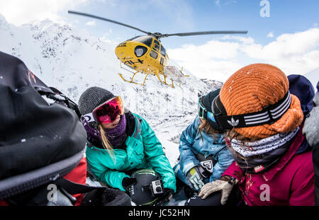 Professional snowboarders Helen Schettini, Jamie Anderson, and Robin Van Gyn, crouch down as a helicopter comes in to land next to them on a sunny blue bird day in Haines, Alaska. Stock Photo