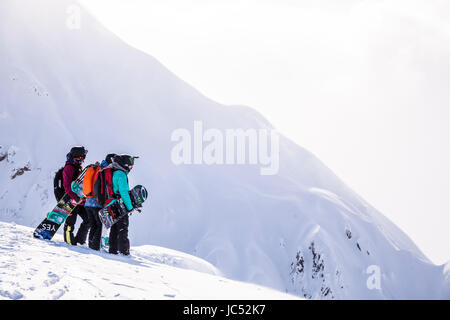 Professional snowboarders Robin Van Gyn, Helen Schettini, and Jamie Anderson, stand on a ridge and look down at a line they are about to ride on a sunny day in Haines, Alaska. Stock Photo