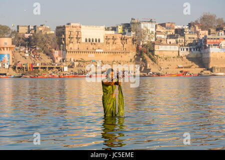 A woman, pilgrim, is taking bath and praying on the sand banks at the holy river Ganges, panorama of Dashashwamedh Ghat, Main Ghat, in the distance Stock Photo