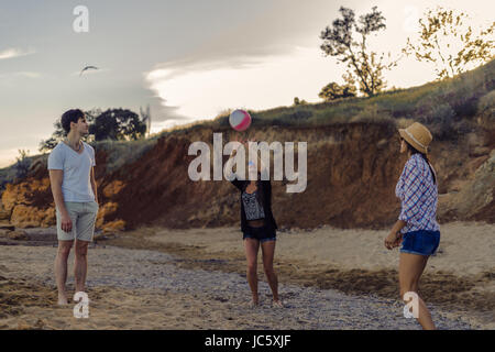 A group of young and cheerful friends playing volleyball on a wild beach during golden sunset. Ball is in air Stock Photo