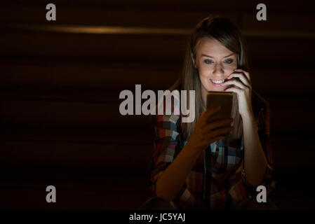 Gorgeous young woman standing with mobile phone at night street outdoors, female caucasian student reading text messages on her cell phone with reflec Stock Photo