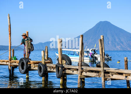 Lake Atitlan, Guatemala - April 4, 2016: Local man dressed in traditional Mayan pants pulls in boat with San Pedro volcano behind. Stock Photo