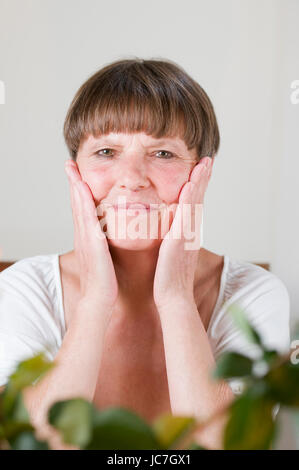 frontales Porträt einer älteren Frau hinter Blumen am Tisch sitzend sich die Hände an die Wangen haltend und in die Kamera lächelnd Stock Photo