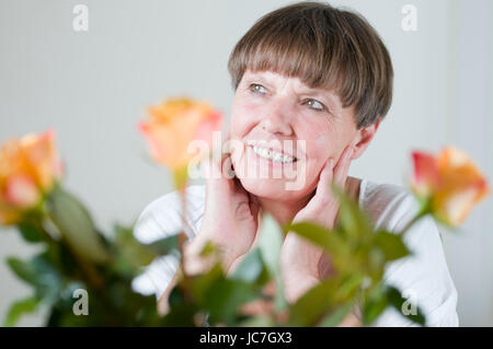frontales Porträt einer älteren Frau hinter Blumen am Tisch sitzend sich die Hände an die Wangen haltend und nach schräg oben links schauend Stock Photo