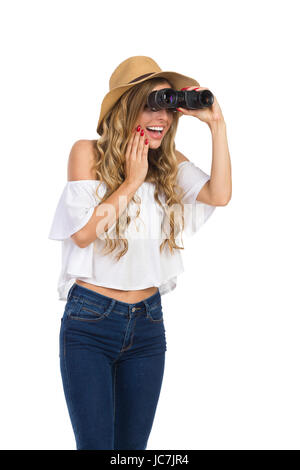 Excited blond young woman in straw hat, jeans and white shirt holding hand on chin looking through binoculars and smiling. Three quarter length studio Stock Photo