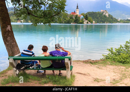 Bled, Slovenia - June 3, 2017: Tourists sitting on a bench on the bank of lake Bled. Church of background Stock Photo