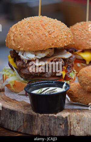 Big American burger with pulled pork meat, onion rings, cheese and salad in sesame bun on paper parchment over natural wooden cut, close up, low angle Stock Photo