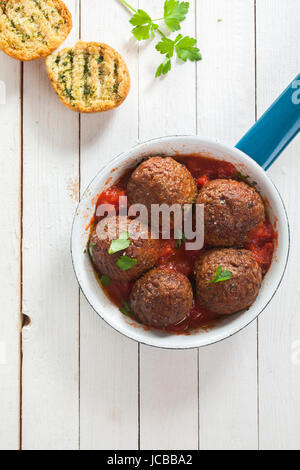 Tasty savory ground beef meatballs in tomato sauce served in a saucepan with crusty toasted herb bread on a white rustic wooden table, overhead view Stock Photo