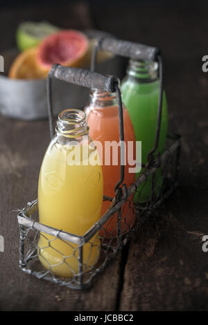 Three full bottles of natural juice, yellow, orange and green, made from squeezed citrus fruits as lemon, grapefruit and green pomelo, in a metallic rustic basket, on a wooden table Stock Photo