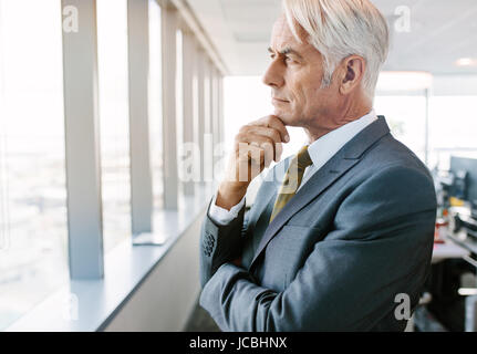 Side view shot of mature businessman standing beside office window looking outside and thinking with hand on chin. Senior professional thinking for so Stock Photo