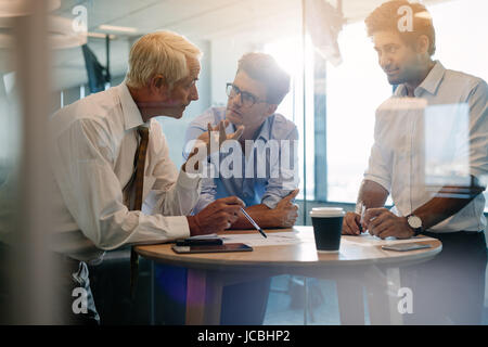 Three male executives standing and discussing around a table. Corporate professional having an informal meeting in modern office. Stock Photo