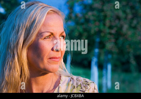 seitliches Kopf-und-Schulter-Porträt einer blonden Frau mittleren Alters in einer Parkanlage in den Sonnenuntergang blickend Stock Photo