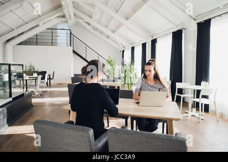 Female business team having a meeting together to discuss paperwork sitting at a table having a discussion. Two businesswomen sitting at a table with laptops and one asking another. Stock Photo