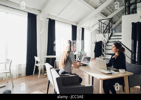 Business women discuss on meeting in modern office. Two female co-workers sitting at the same table with laptops. Stock Photo
