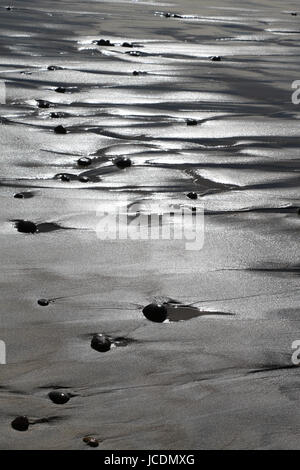 Newly exposed sandy beach glows in the sun as the tide recedes,leaving rocks and water channels, and a silvery reflective light on the sand. Stock Photo