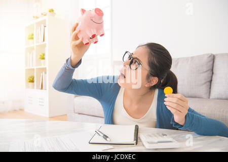 young unhappy woman emptying her piggybank savings with less than expected sitting on sofa in the living room at home. interior and domestic housework Stock Photo