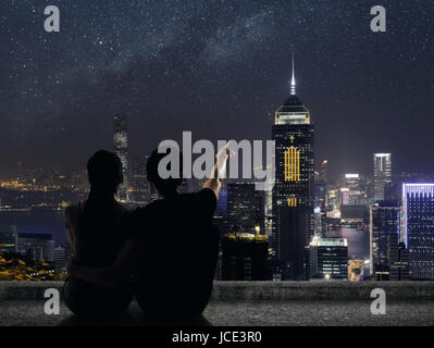 Silhouette of couple sit on ground point faraway on the roof above the city in the night. Stock Photo