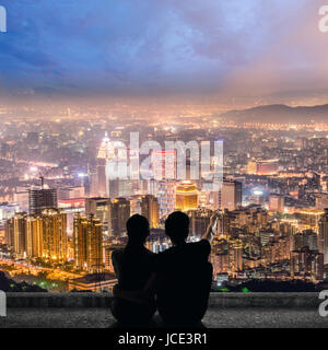 Silhouette of couple sit on ground point faraway on the roof above the city in the night. Stock Photo