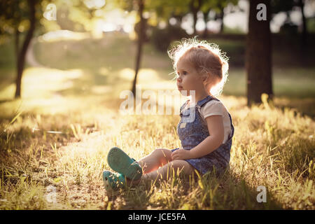 Kid rest in park in the evening. Beautiful little girl in summer park sits on grass. Retro style. Stock Photo