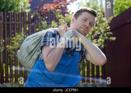 Caucasian gardener in overalls and working gloves carrying bag with garbage in summer day Stock Photo