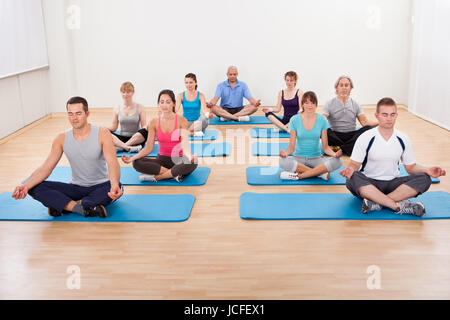 Diverse group of people practicing yoga in a gym sitting cross-legged on their mats meditating Stock Photo