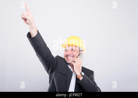 Architect smiling pointing up and talking on smartphone wearing yellow hardhat on grey background Stock Photo