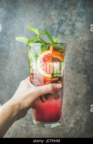 Woman's hand holding glass of blood orange citrus lemonade with mint and ice, dark brown stone background. Refreshing summer drink concept Stock Photo