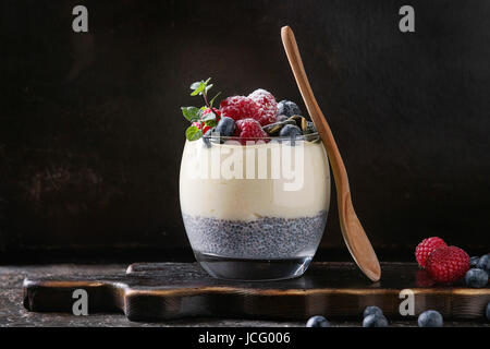 Dessert breakfast layered chia seeds pudding, rice porridge in glass decorated by fresh blueberries, raspberry, mint. Stand with wooden spoon on dark