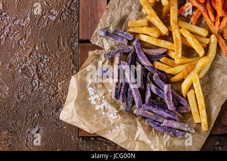Variety of french fries traditional potatoes, purple potato, carrot served with salt on baking paper over brown texture background. Top view with spac Stock Photo